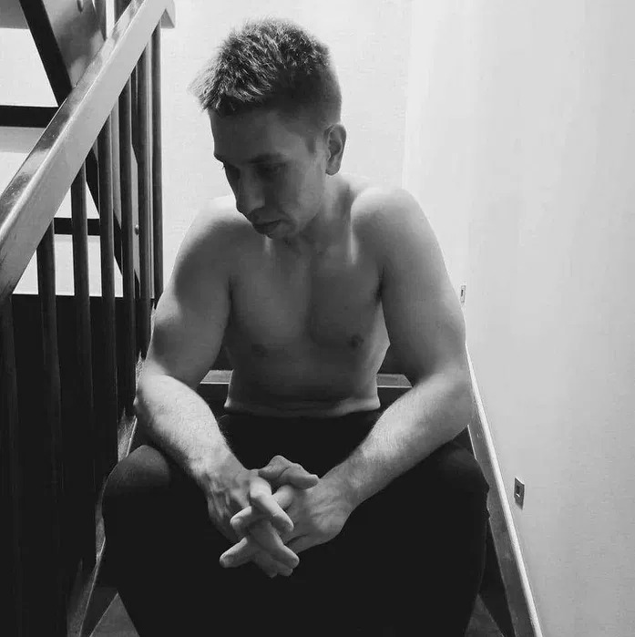 Mężczyzna bez koszulki siedzi na schodach, zamyślony, z dłońmi splecionymi na kolanach, zdjęcie w czerni i bieli.