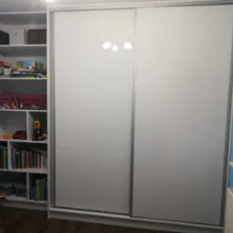 Biała szafa wnękowa z przesuwnymi drzwiami i regał z półkami wypełniony książkami i zabawkami w pokoju dziecięcym, widoczne odbicie lampy na drzwiach szafy.