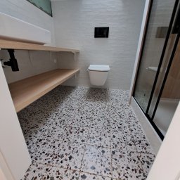 Przemysław Proczek - Nowoczesna łazienka z białą ceglaną ścianą, wiszącą toaletą, kabiną prysznicową z czarnymi ramami i podłogą w terazzo. Półki z jasnego drewna zamontowane pod umywalką.