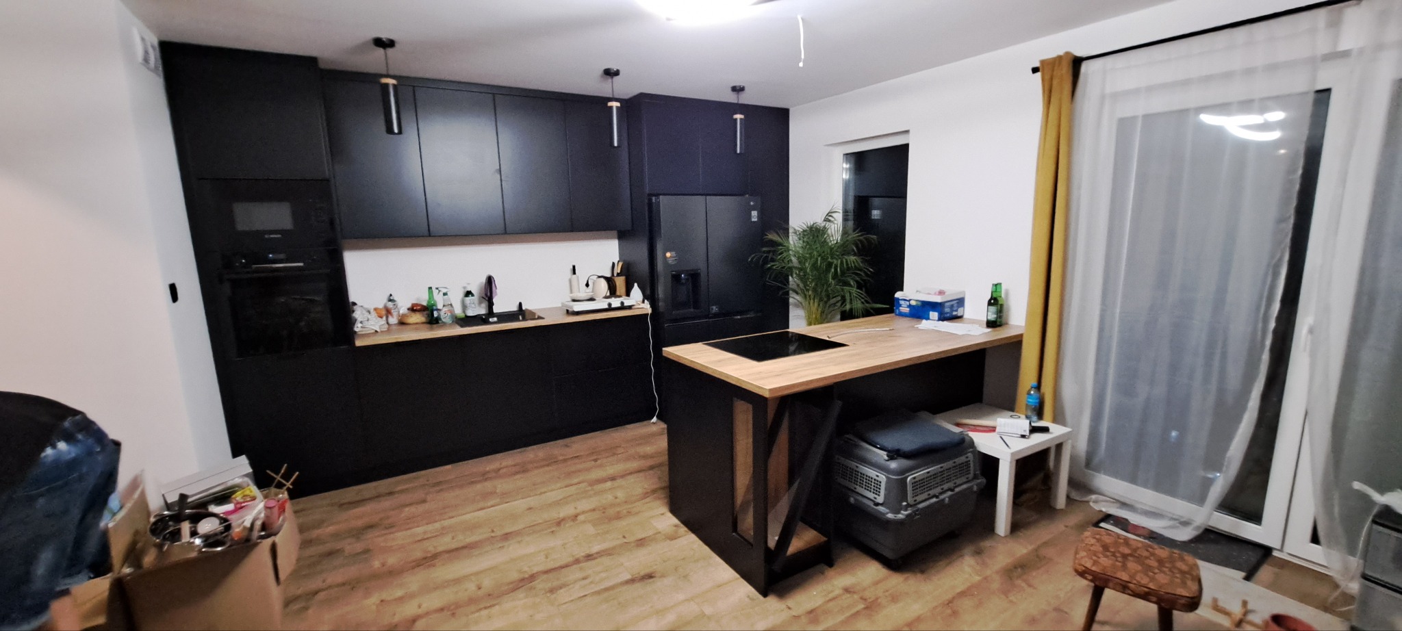 Nowoczesna kuchnia z czarnymi szafkami i drewnianym blatem wyspy, czarne lampy wiszące, widoczny fragment kartonu z akcesoriami kuchennymi w rogu, nie do końca posprzątane otoczenie.