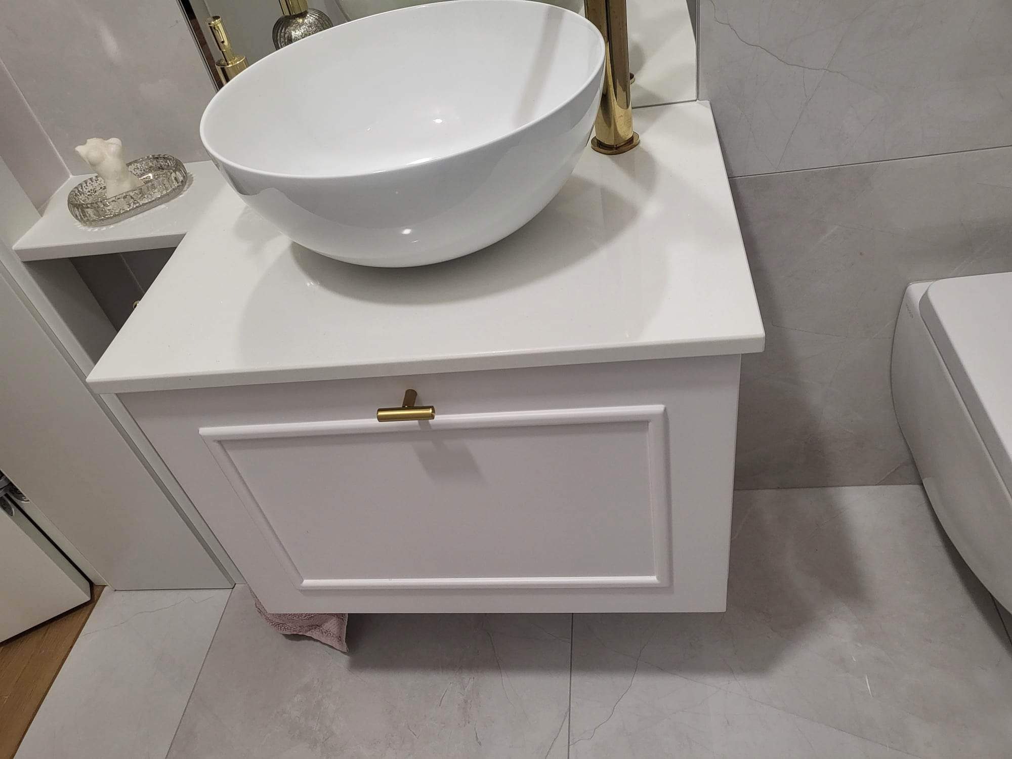 Biała, okrągła umywalka nablatowa z złotą armaturą, osadzona na białej szafce z frezowanymi ramkami i złotym uchwytem, na tle szarych płytek imitujących marmur w wykończonej łazience.