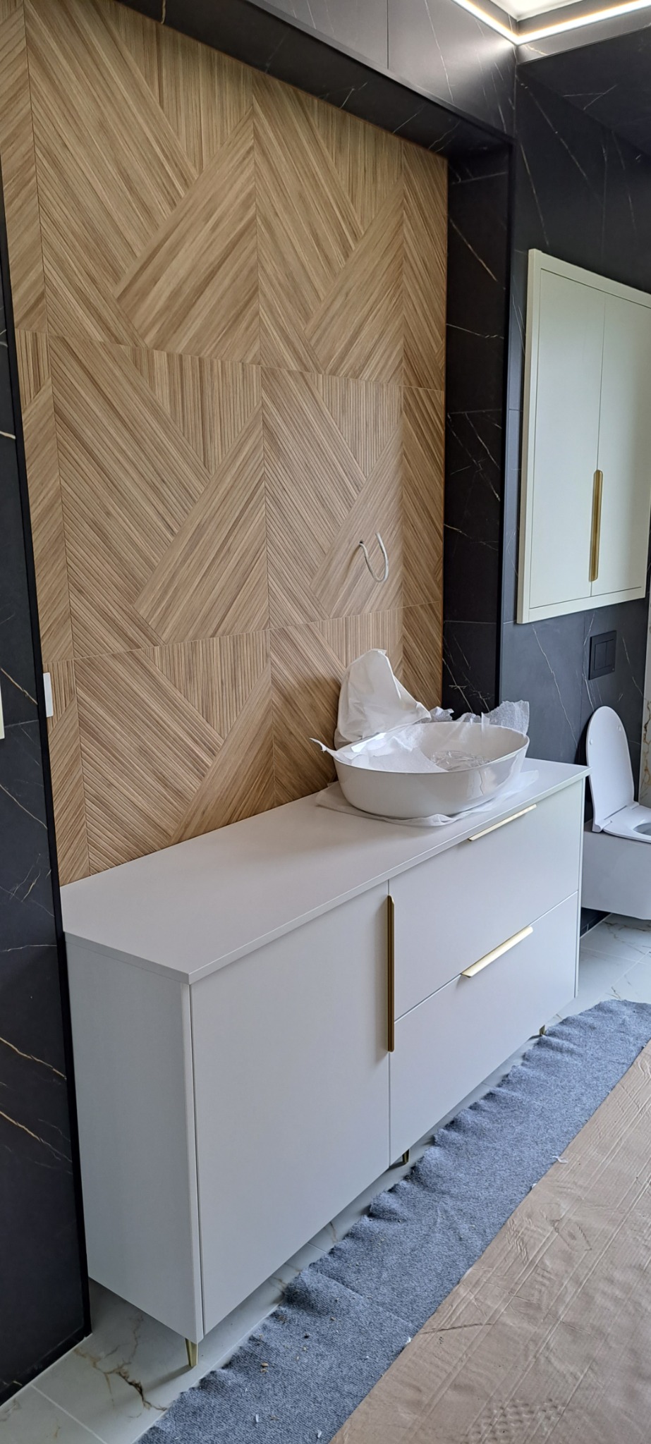 Nowoczesna łazienka w Segnowach z białą szafką pod umywalkę z złotymi uchwytami, drewnianą mozaiką na ścianie i czarnymi marmurowymi płytkami, w trakcie montażu umywalki.