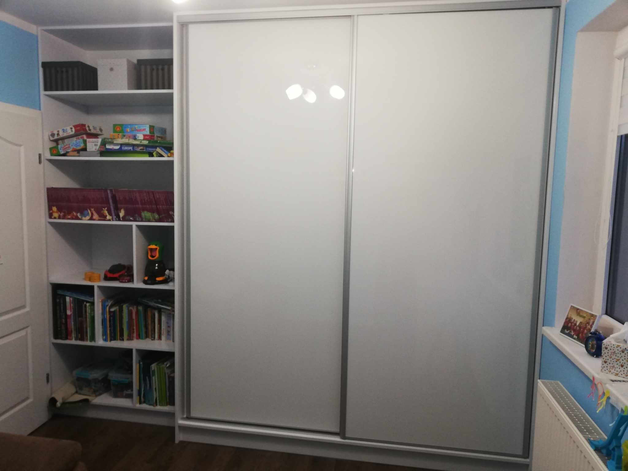 Biała szafa wnękowa z przesuwnymi drzwiami i regał z półkami wypełniony książkami i zabawkami w pokoju dziecięcym, widoczne odbicie lampy na drzwiach szafy.