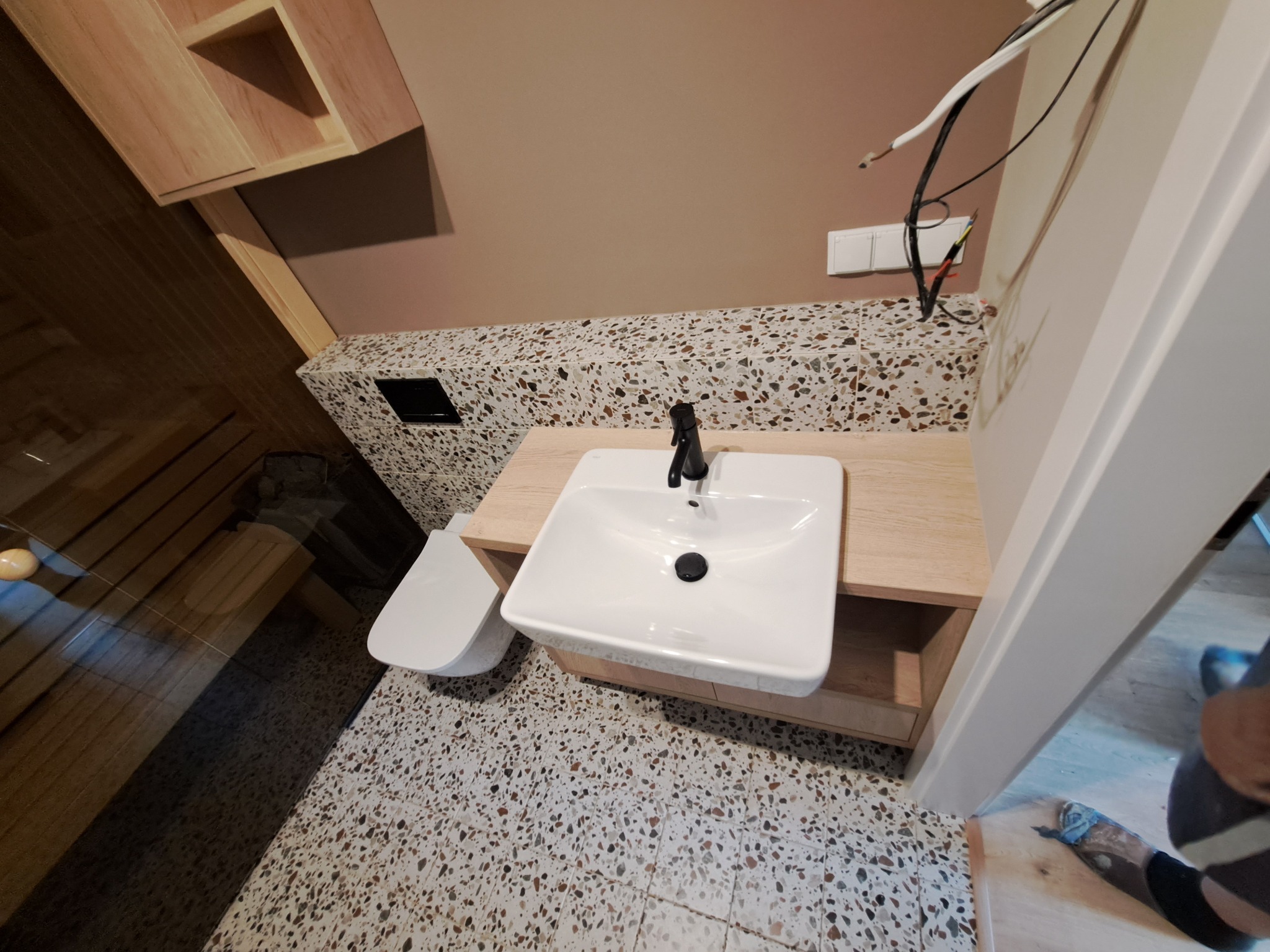 Nowoczesna łazienka z umywalką nablatową, toaletą wiszącą i mozaikową podłogą, widoczne kable elektryczne i fragment sauny.