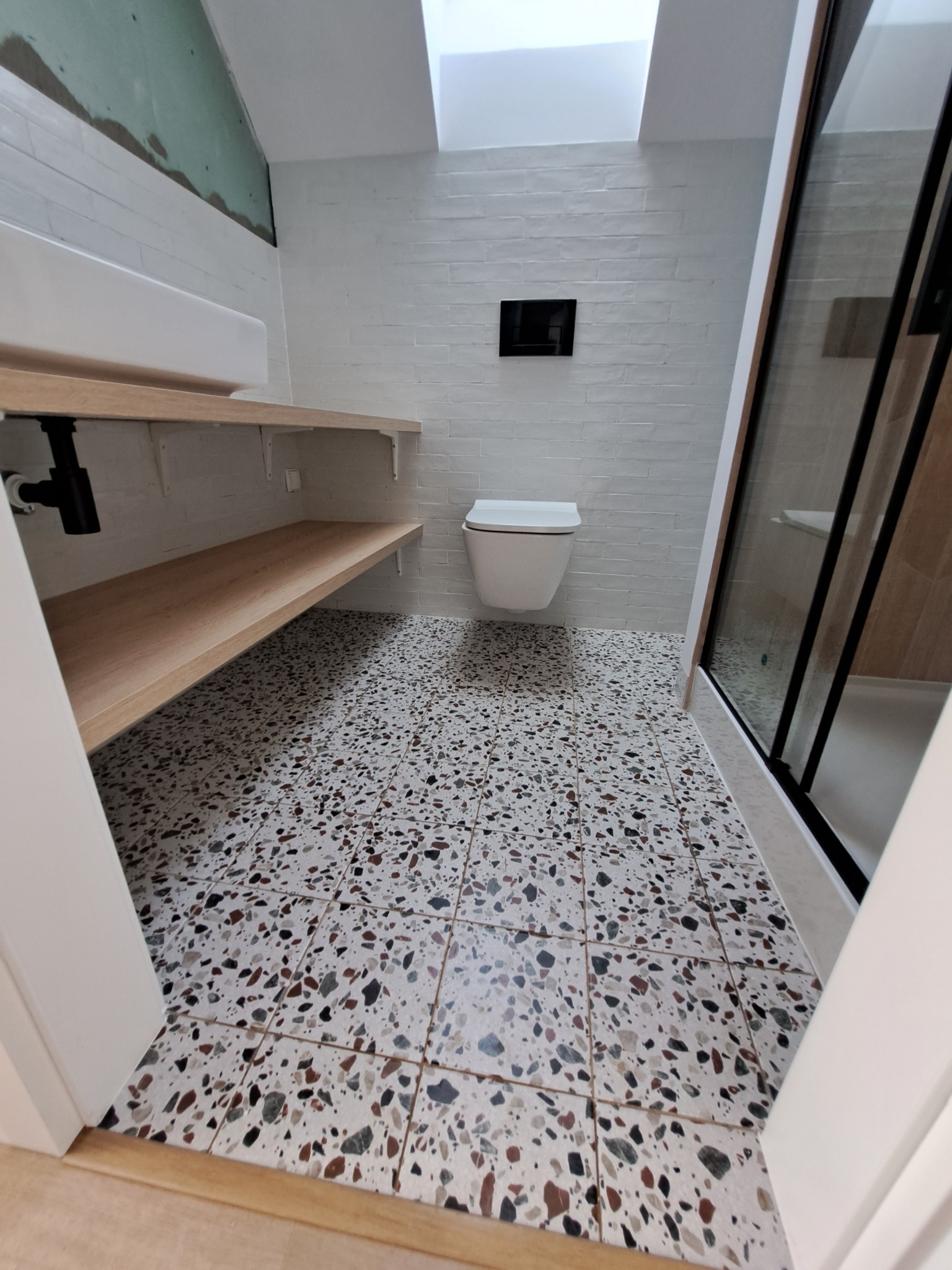 Nowoczesna łazienka z białą ceglaną ścianą, wiszącą toaletą, kabiną prysznicową z czarnymi ramami i podłogą w terazzo. Półki z jasnego drewna zamontowane pod umywalką.