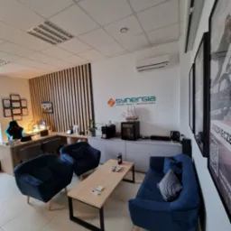 Wnętrze biura firmy Synergia z logo na ścianie, oferującej leasing, kredyty i ubezpieczenia. Wygodne, granatowe fotele i sofa, stolik kawowy oraz biurko z certyfikatami w tle tworzą przyjazną...