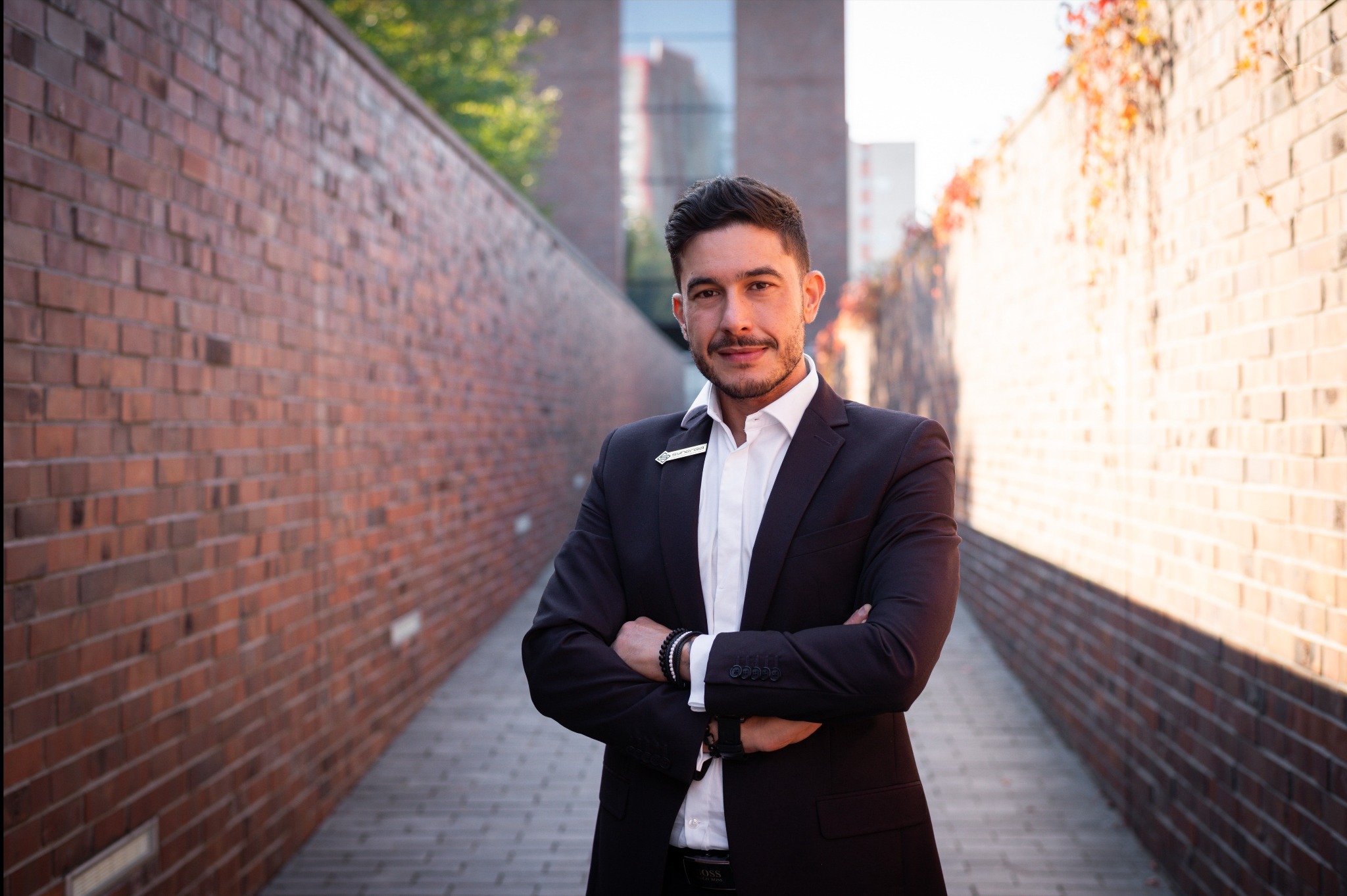 Mężczyzna w garniturze, z rękami skrzyżowanymi na tle ceglanej ściany i budynku. Widoczna plakietka z imieniem. Uśmiechnięty, pewny siebie.
