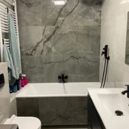 Nowoczesna łazienka z szarymi płytkami imitującymi marmur, czarną armaturą i białym wyposażeniem, w tym grzejnik drabinkowy z powieszonymi ręcznikami w kolorze morskim.