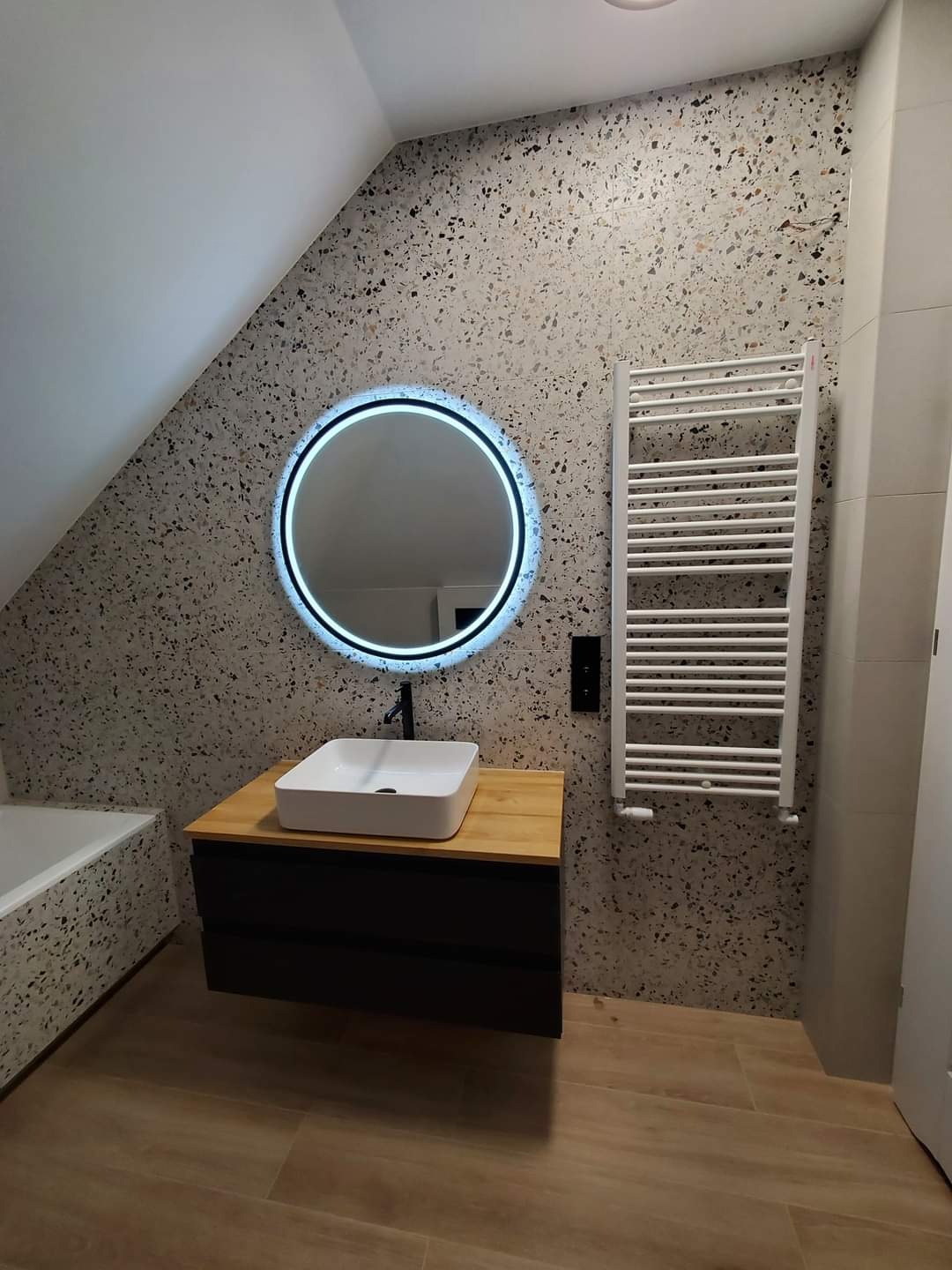 Nowoczesna łazienka na poddaszu z umywalką nablatową, podświetlanym okrągłym lustrem i grzejnikiem drabinkowym na ścianie wykończonej płytkami lastryko.