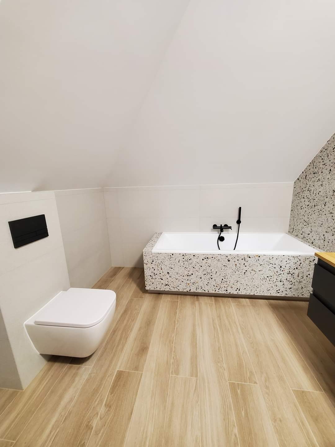 Nowoczesna łazienka na poddaszu z drewnopodobną podłogą, wiszącą toaletą, białą wanną obudowaną płytkami lastryko i czarną armaturą.