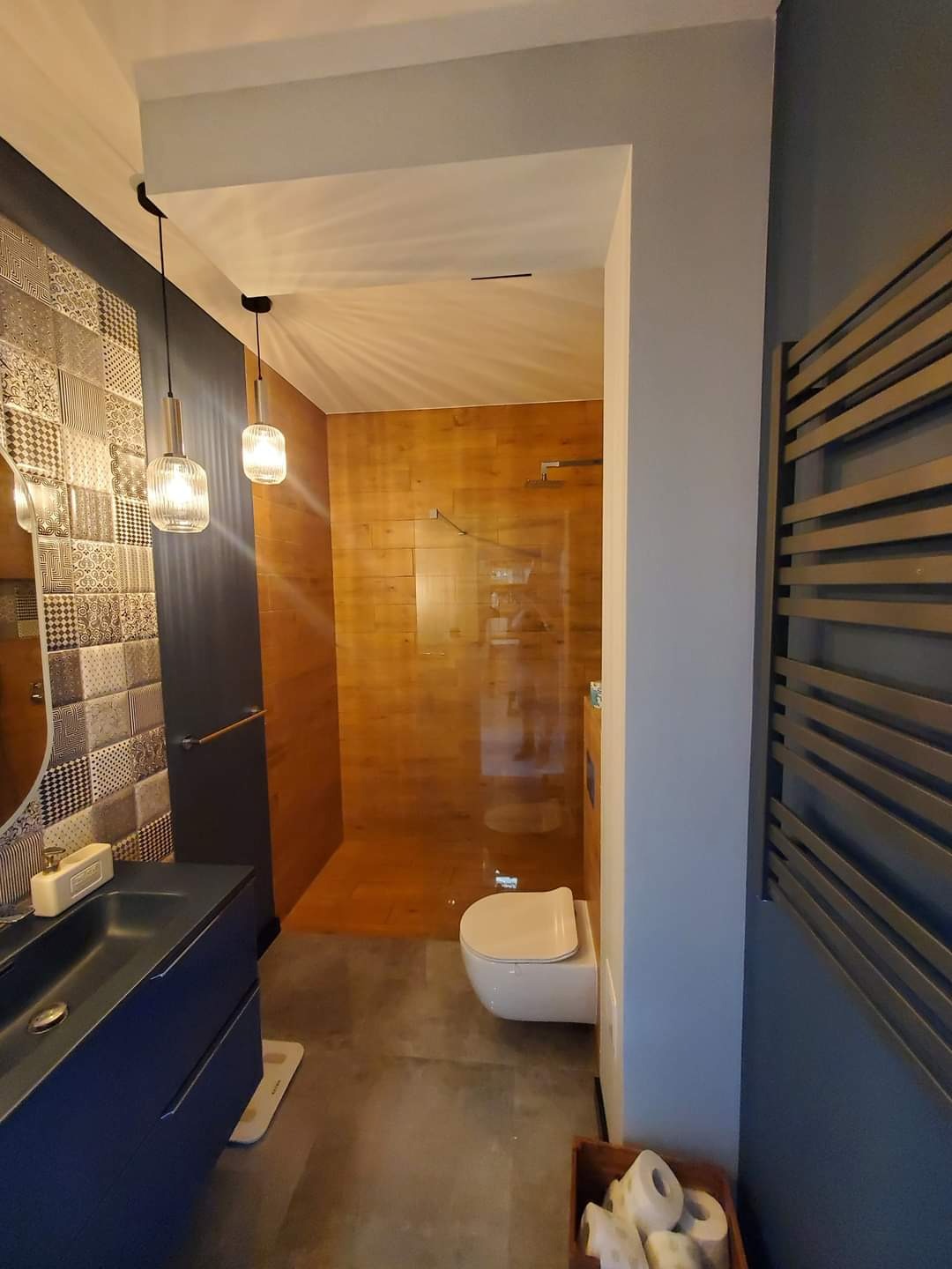 Nowoczesna łazienka z drewnopodobnymi płytkami w kabinie prysznicowej, wiszącą toaletą, ciemnoniebieską szafką z umywalką i dekoracyjnymi płytkami nad nią, oraz grzejnikiem drabinkowym w kolorze...