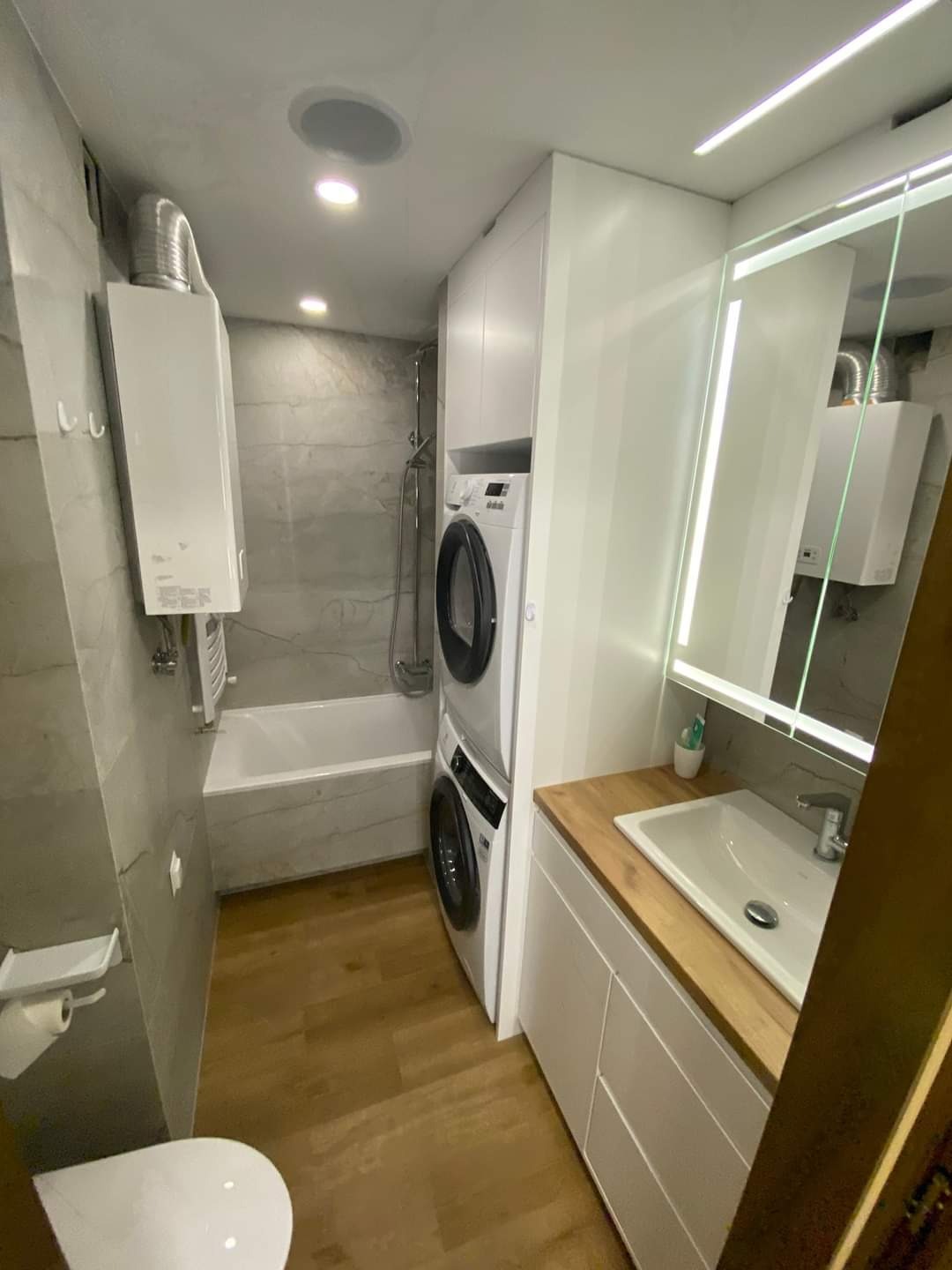 Nowoczesna łazienka z wanną, pralko-suszarką w zabudowie, umywalką na drewnianym blacie i szarą płytką imitującą marmur na ścianach.