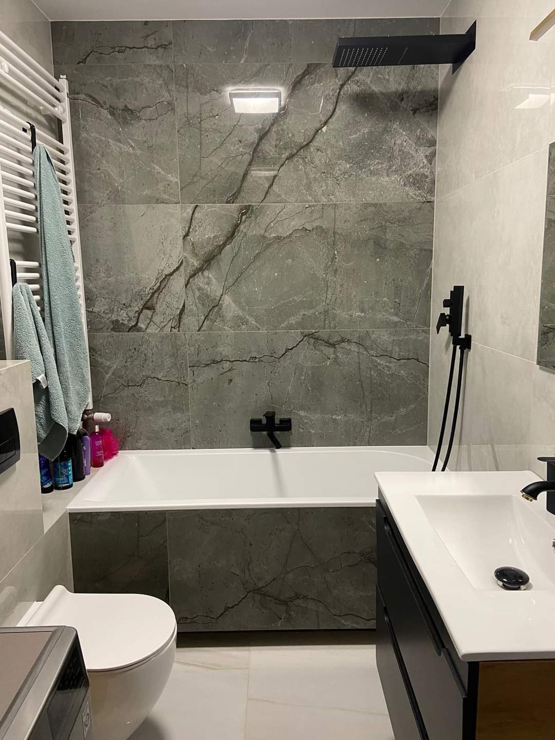 Nowoczesna łazienka z szarymi płytkami imitującymi marmur, czarną armaturą i białym wyposażeniem, w tym grzejnik drabinkowy z powieszonymi ręcznikami w kolorze morskim.