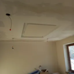 Niedokończony sufit z gładzią, z widocznym obniżeniem w kształcie kwadratu, wystającymi przewodami elektrycznymi oraz fragmentem okna z widokiem na zimowy krajobraz.