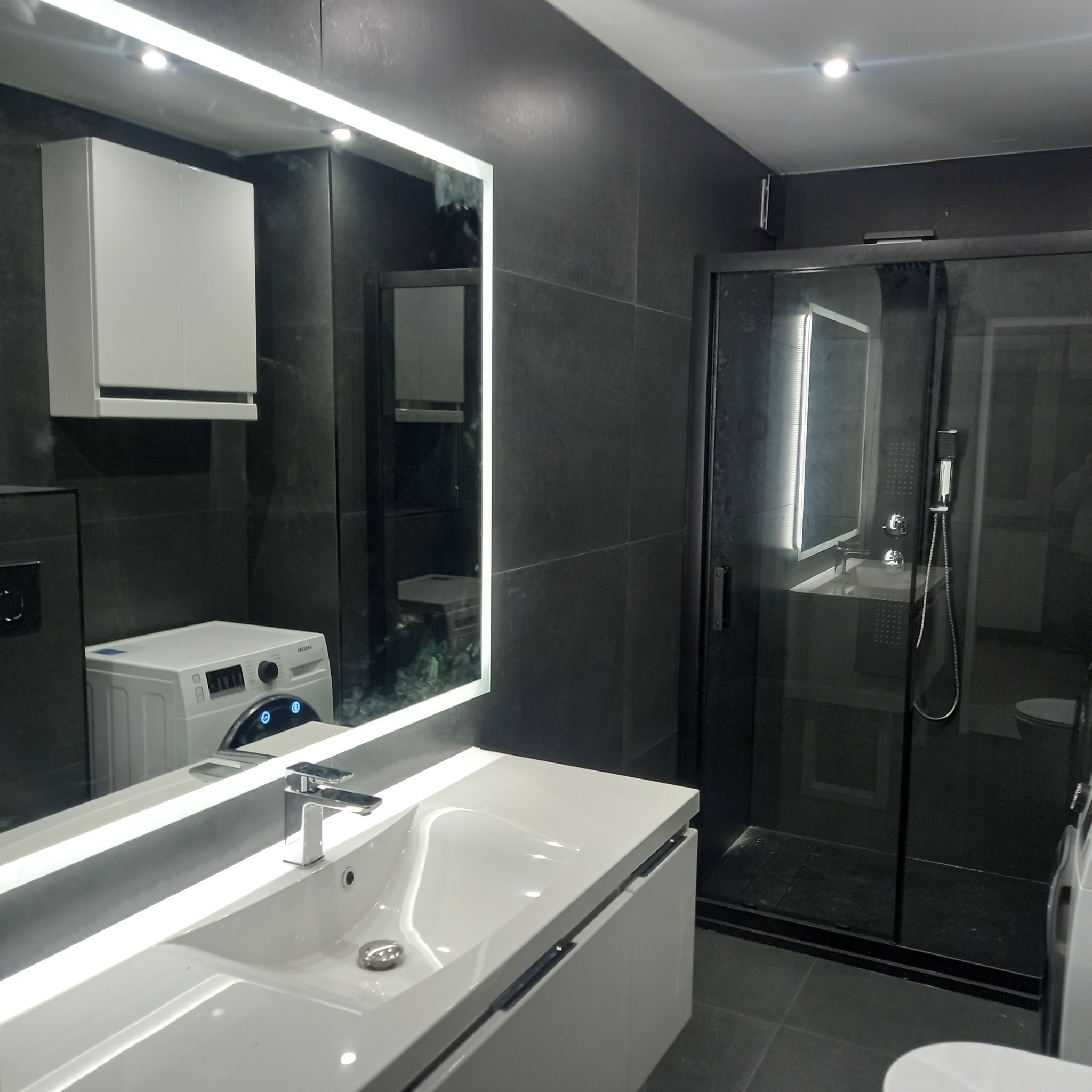 Nowoczesna łazienka z czarnymi płytkami, podświetlanym lustrem nad białą umywalką z szafką, kabiną prysznicową i pralką w tle.