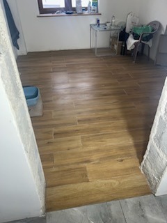 Ułożona podłoga z płytek imitujących drewno w pomieszczeniu w trakcie remontu, widoczne niedoskonałości i pozostałości po pracach budowlanych, w tle okno i stół z materiałami budowlanymi.