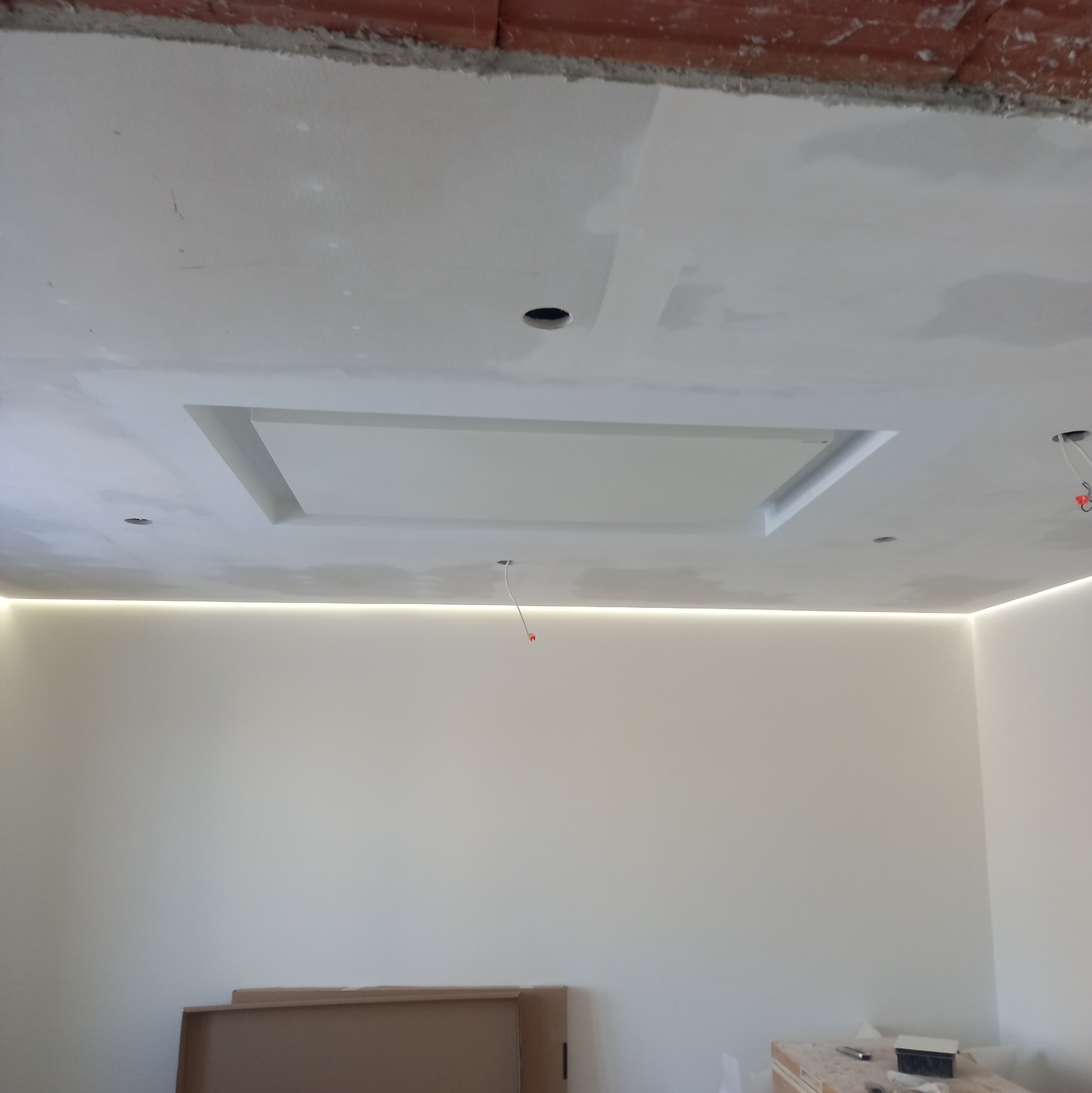 Wnętrze pokoju w trakcie prac wykończeniowych: sufit z wnęką podświetloną ledami, ściany po szpachlowaniu, widoczne kable wystające z sufitu, kartony na podłodze.