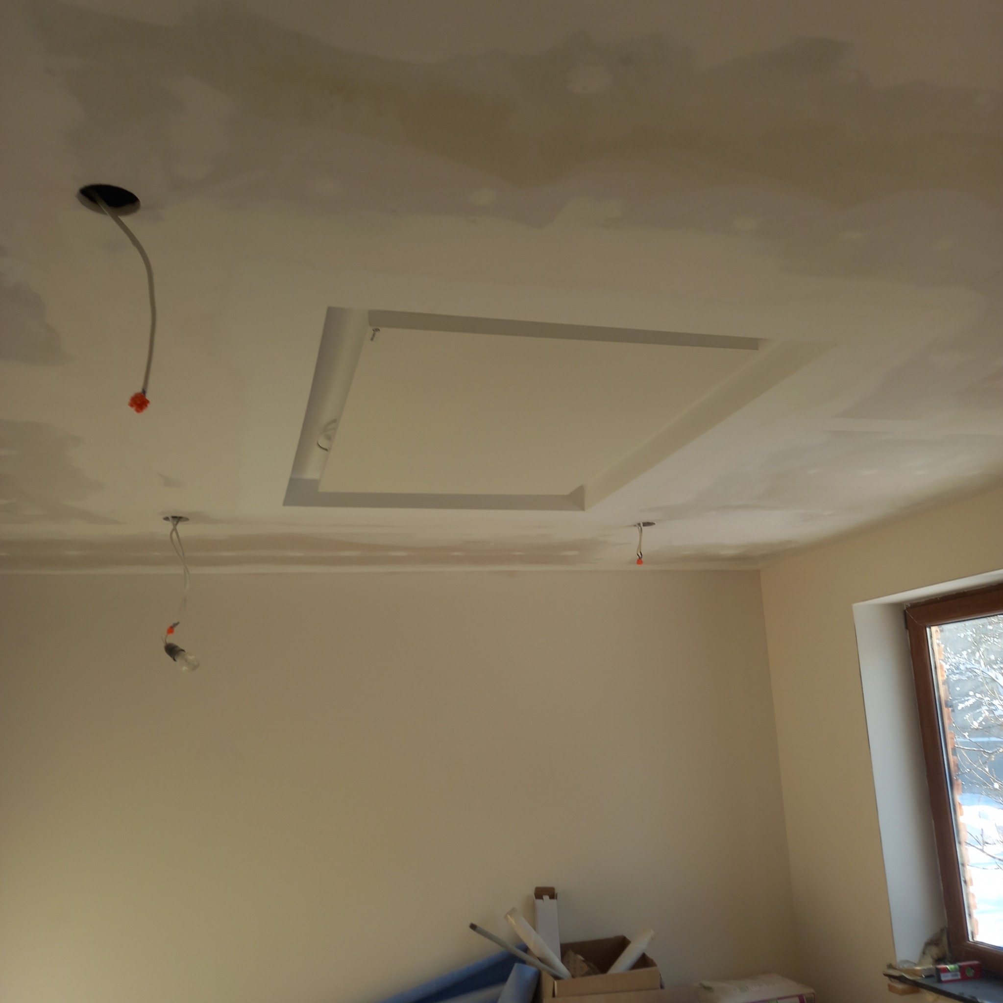 Niedokończony sufit z gładzią, z widocznym obniżeniem w kształcie kwadratu, wystającymi przewodami elektrycznymi oraz fragmentem okna z widokiem na zimowy krajobraz.