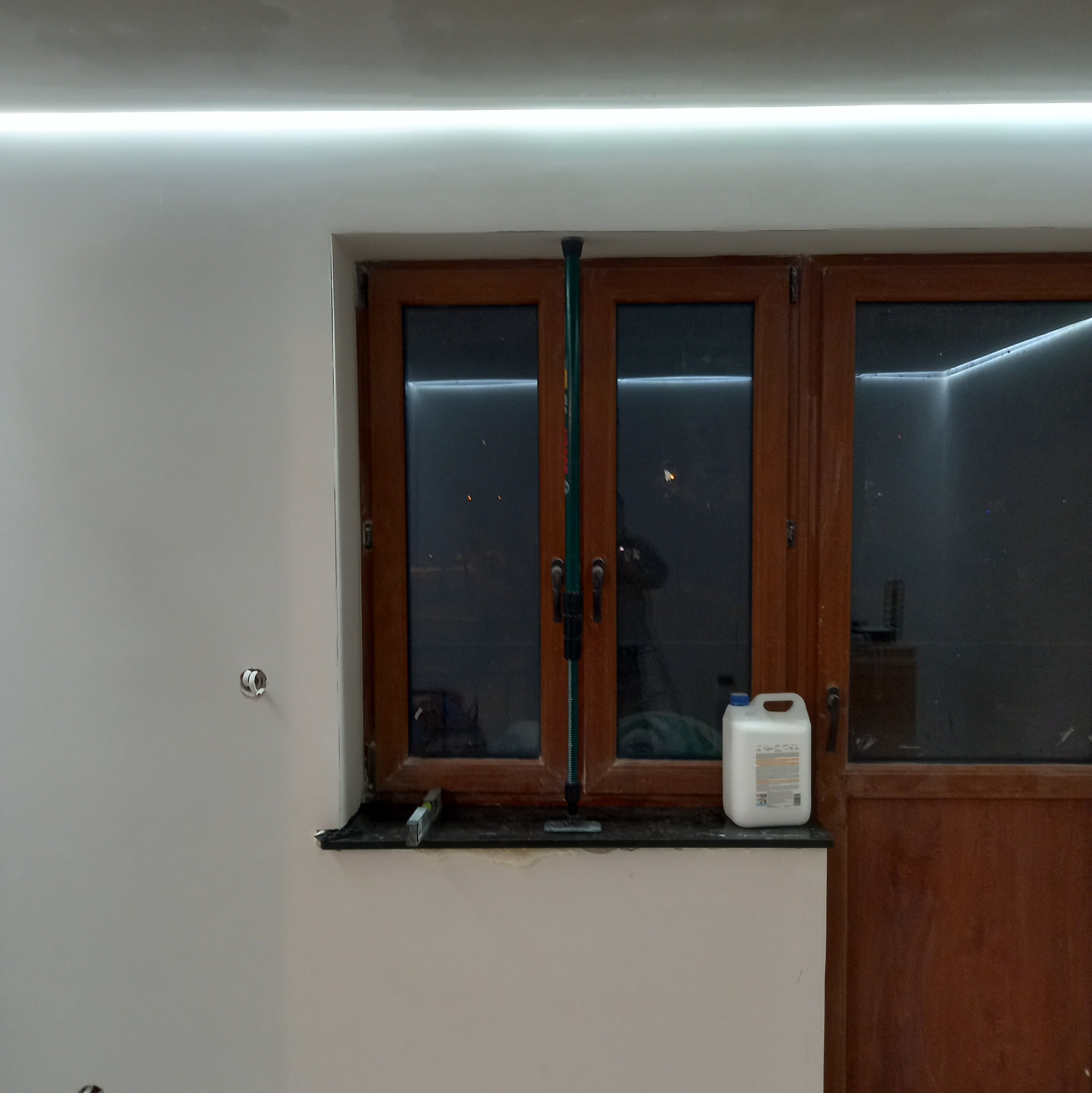 Świeżo wykończona ściana w Bielsko-Białej z zamontowanym oświetleniem LED wzdłuż sufitu, drewniane okno z poziomnicą i kanistrem farby na parapecie.