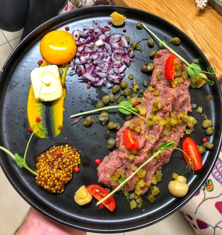 Czarny talerz z tatarem wołowym, żółtkiem, czerwoną cebulą, kaparami, musztardą francuską i dekoracjami z mikro ziół, trzymany w dłoni.