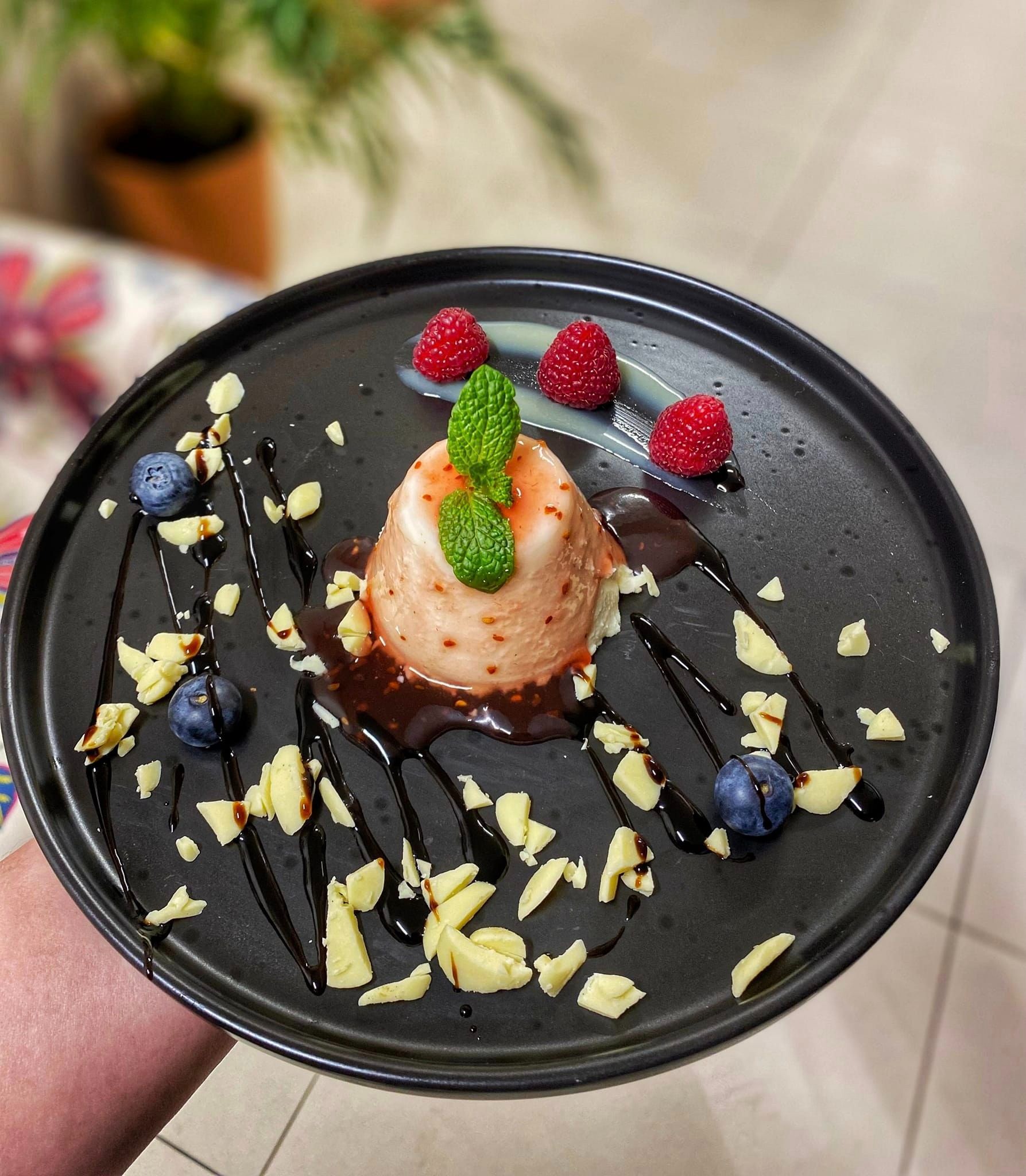 Elegancko podane Panna Cotta na czarnym talerzu, udekorowane malinami, borówkami, listkami mięty, kawałkami białej czekolady i sosem czekoladowym, z rośliną w tle.