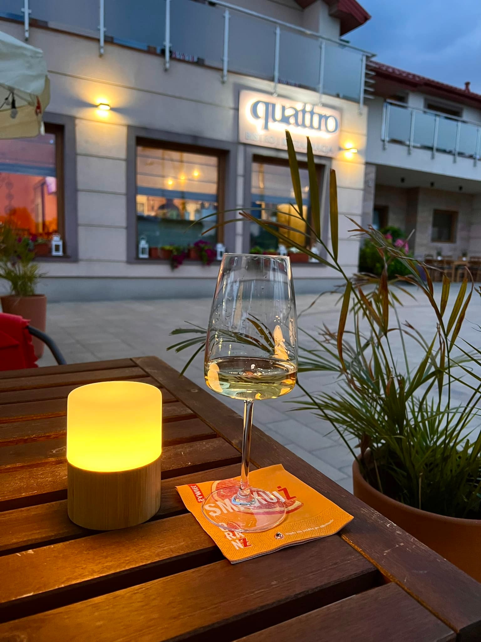 Stół na zewnątrz restauracji o zmierzchu, z lampką, kieliszkiem białego wina i serwetką reklamową. W tle budynek z napisem 'quattro'.