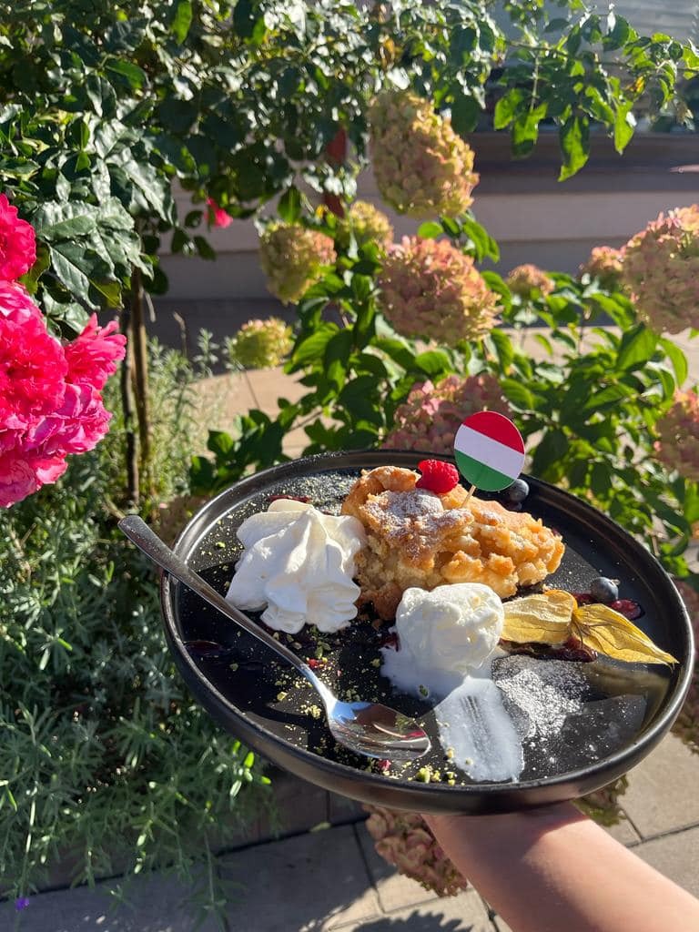 Jabłecznik z bitą śmietaną i lodami na czarnym talerzu, udekorowany flagą Węgier, podany na tle kwitnących krzewów.