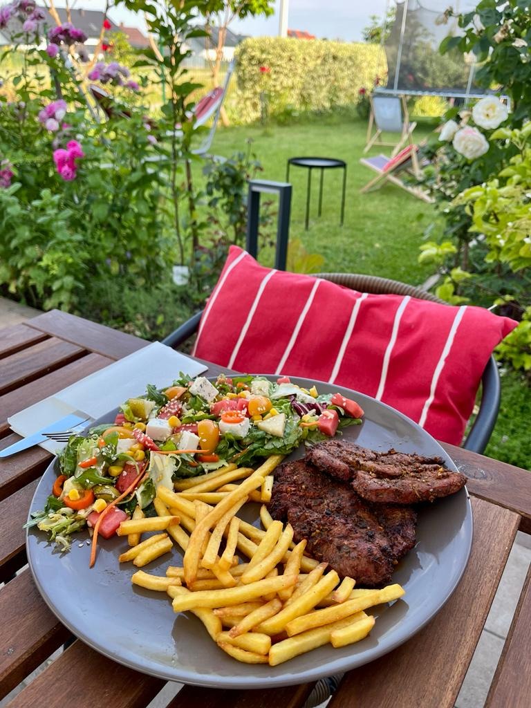 Stek z frytkami i sałatką na talerzu, na drewnianym stole w ogrodzie z kwitnącymi różami i leżakami w tle.