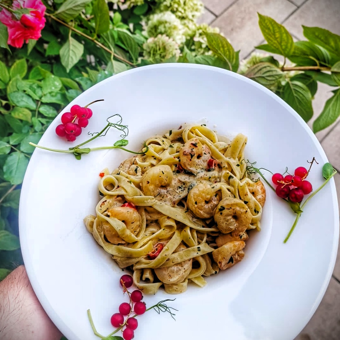 Elegancko podane tagliatelle z krewetkami na białym talerzu, ozdobione gałązkami czerwonych jagód i zielenią, na tle bujnej roślinności.