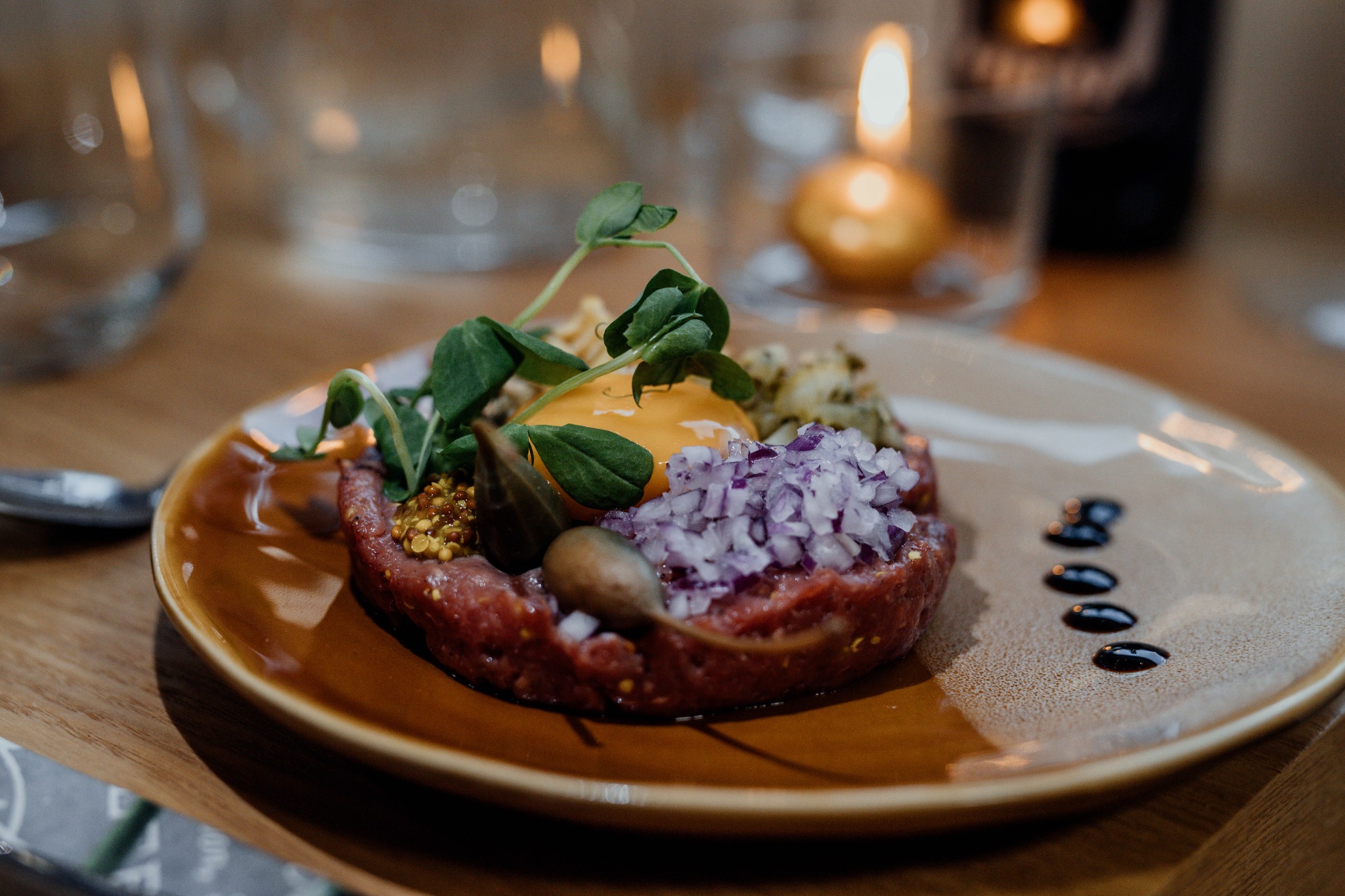 Tatar wołowy udekorowany czerwoną cebulą, musztardą francuską, kaparami, żółtkiem i mikro-ziołami, podany na brązowym talerzu z kroplami sosu balsamicznego w tle ciepłe światło świecy