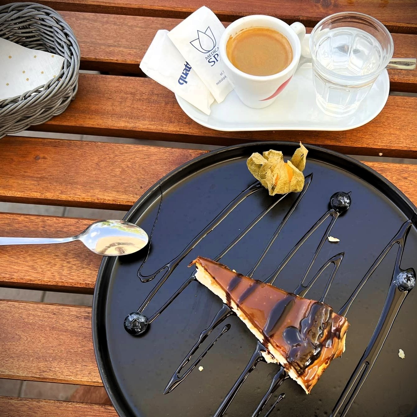 Kawa, woda, sernik z polewą karmelową i physalis na czarnym talerzu z dekoracją z polewy, widok z góry na drewnianym stole.