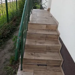 Schody wejściowe wyłożone płytkami imitującymi drewno, prowadzące do drzwi, z zieloną metalową balustradą po lewej stronie i fragmentem elewacji budynku po prawej.