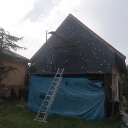 Dom w trakcie ocieplania elewacji: czarna warstwa izolacyjna z białymi kołkami, rusztowanie i drabina oparta o ścianę, przykryte plandeką elementy budynku.