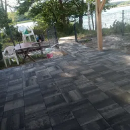 Ułożona, szara kostka brukowa na patio z widokiem na jezioro, z widocznymi krzesłami i stolikiem oraz ogrodzeniem z siatki.