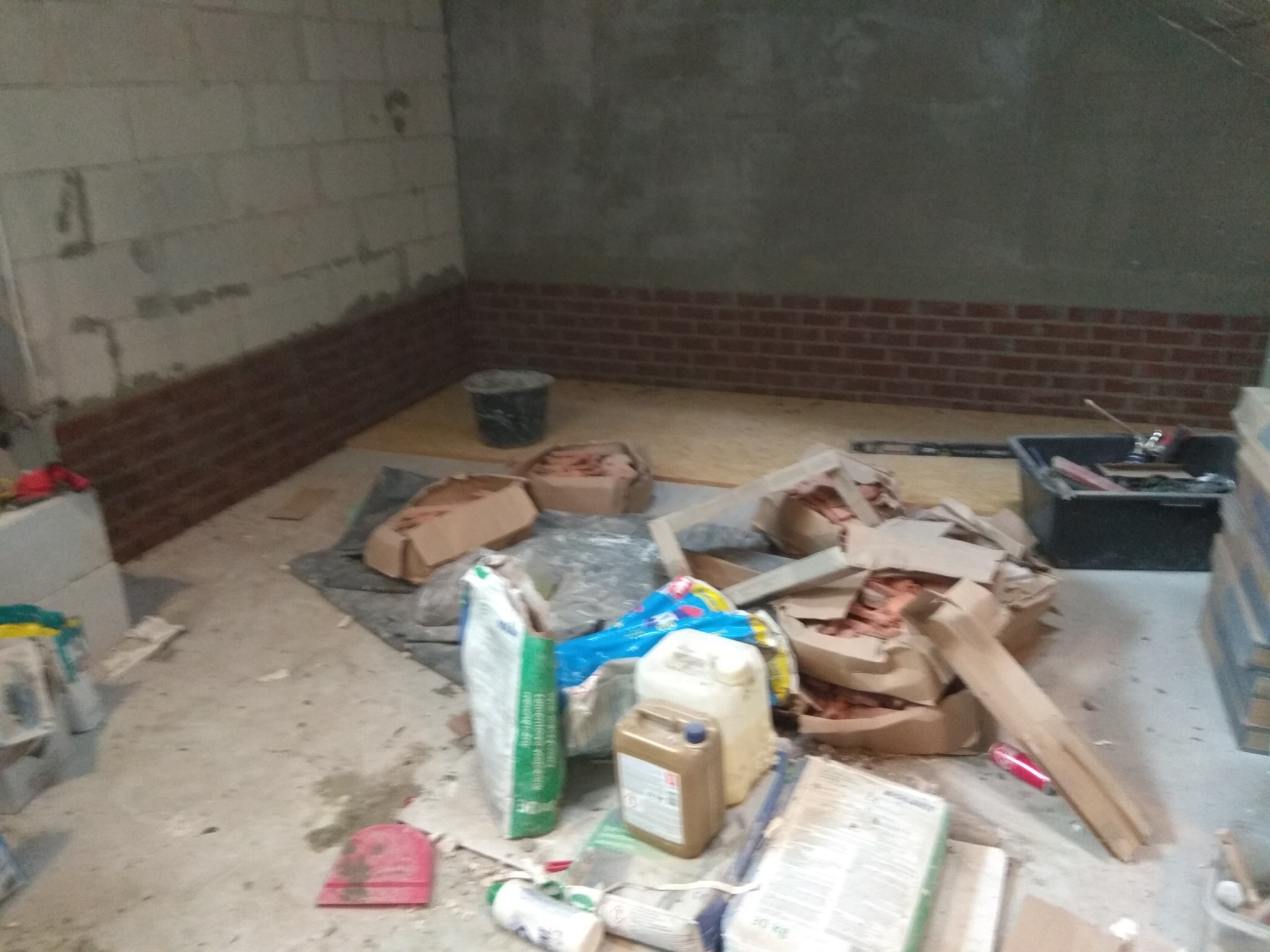 Fragment ściany z bloczków betonowych z częściowo ułożoną dekoracyjną cegłą klinkierową, widoczne materiały budowlane i narzędzia w trakcie prac wykończeniowych.