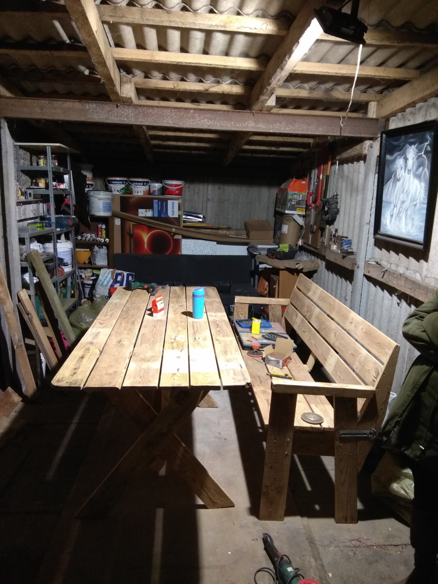 Drewniany stół i ławka w warsztacie, widoczne narzędzia, puszki z farbami i regał z różnymi przedmiotami.