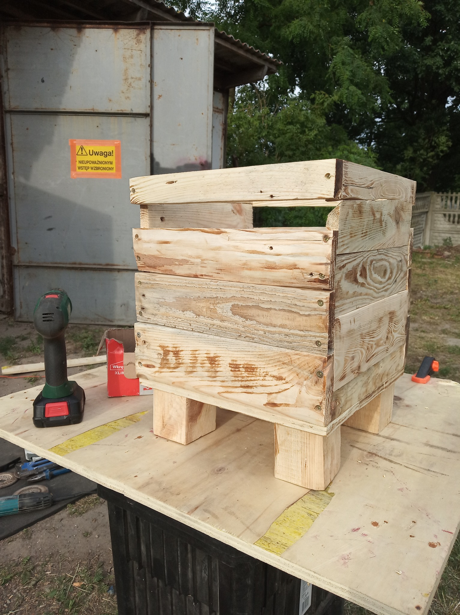 Drewniana skrzynka z desek, skręcona śrubami, na czterech nóżkach, widoczna wiertarka, pudełko po wkrętach i narzędzia na stole warsztatowym w plenerze.