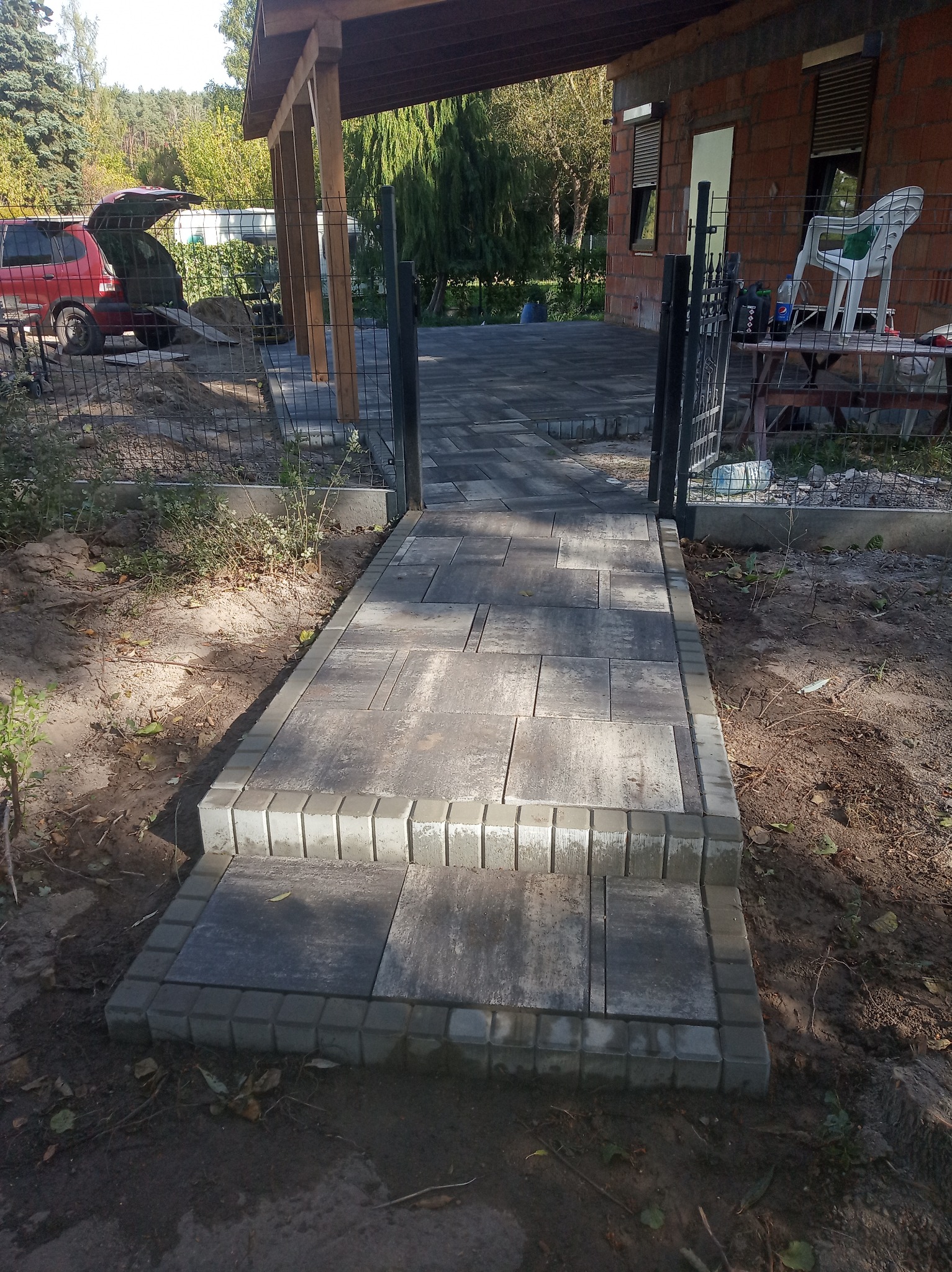 Ułożona ścieżka z kostki brukowej prowadząca do domu z cegły, widoczny samochód z otwartym bagażnikiem i teren w trakcie prac wykończeniowych.