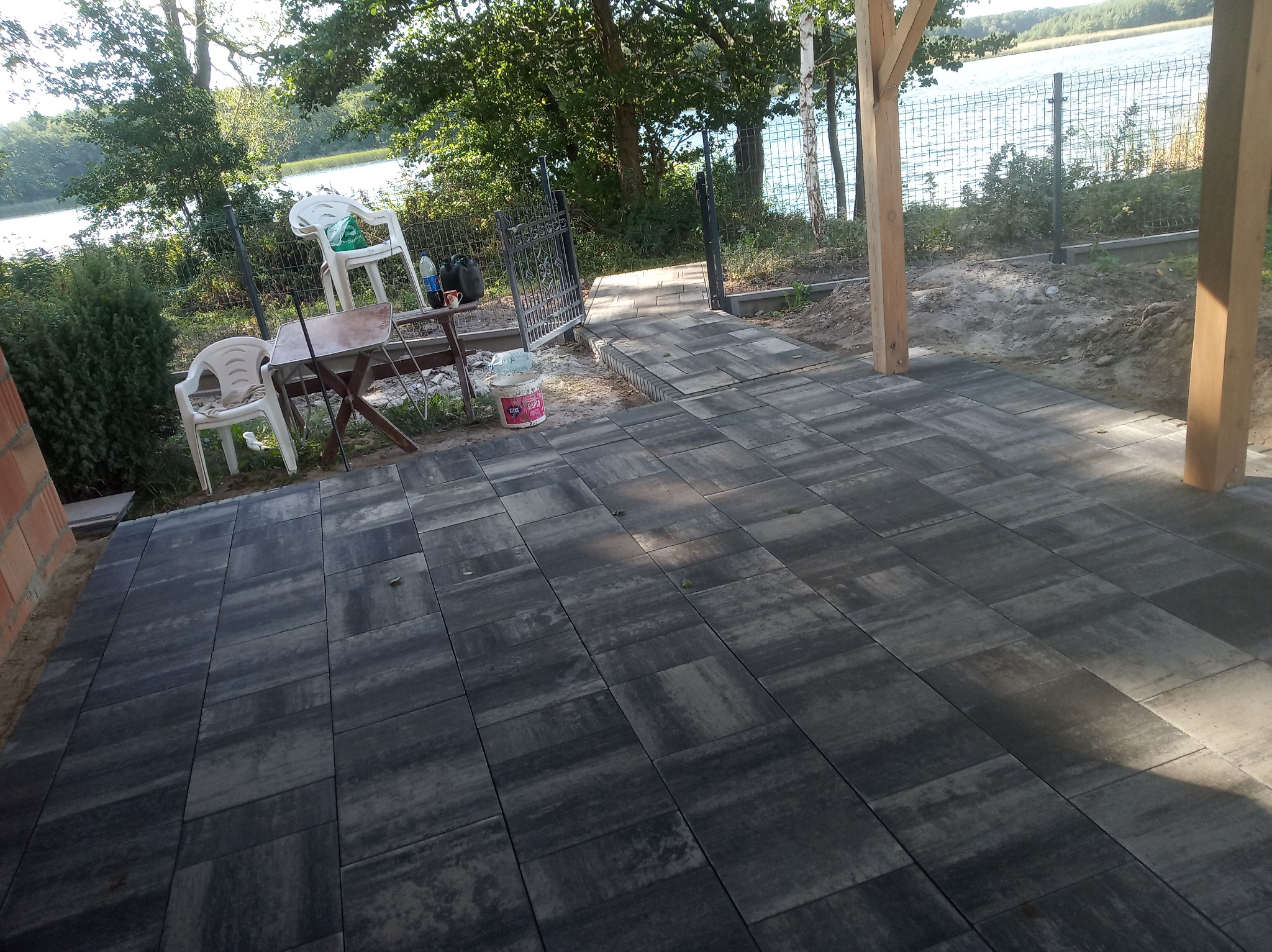 Ułożona, szara kostka brukowa na patio z widokiem na jezioro, z widocznymi krzesłami i stolikiem oraz ogrodzeniem z siatki.