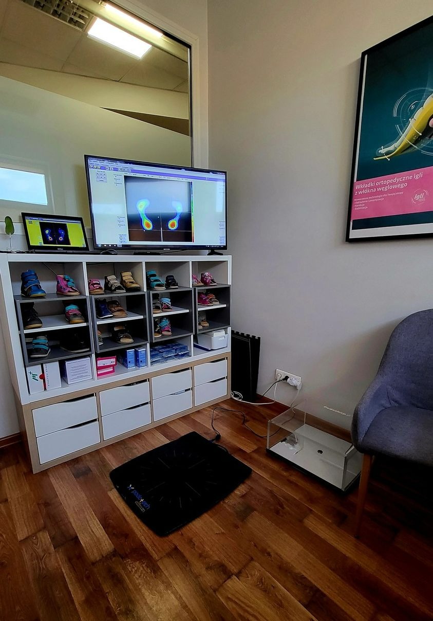 Gabinet z platformą do badania stóp, analizą komputerową rozkładu nacisku, półkami z obuwiem ortopedycznym i plakatem informacyjnym o wkładkach ortopedycznych.