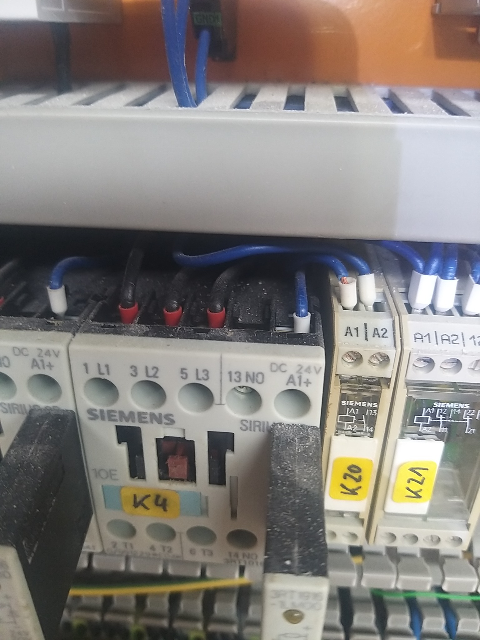 Szczegółowe ujęcie na panel sterowniczy z przekaźnikami Siemens, okablowaniem w niebieskiej izolacji i żółtymi etykietami identyfikacyjnymi K4, K20, K21, widoczne ślady kurzu na elementach.