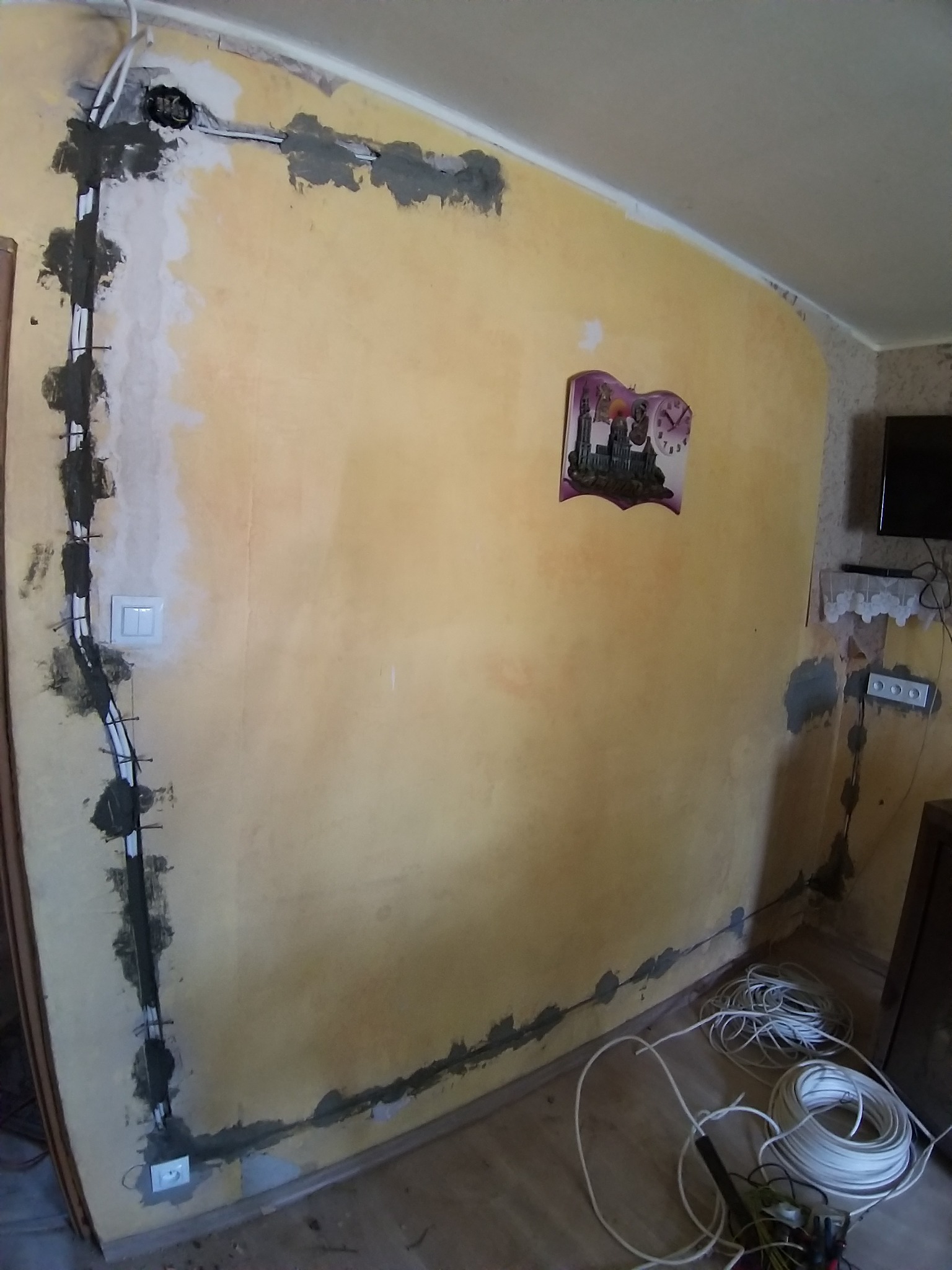 Wewnętrzne kable elektryczne prowadzone po ścianie, mocowane zaprawą gipsową, widoczne puszki instalacyjne, włącznik światła i zwój kabli na podłodze.