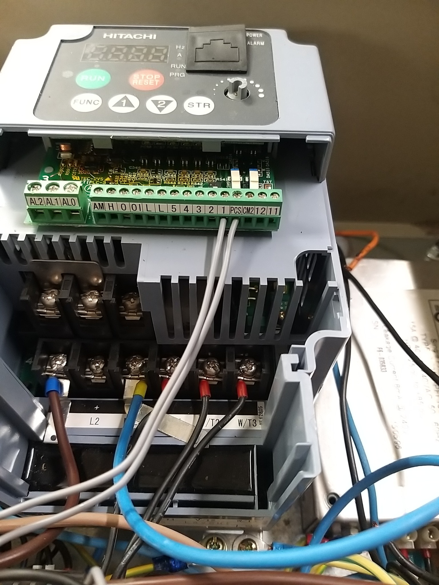 Szary falownik Hitachi z widocznymi zaciskami, podłączonymi przewodami w kolorach niebieskim, brązowym i czarnym, oraz zielonymi listwami z numerowanymi złączami.