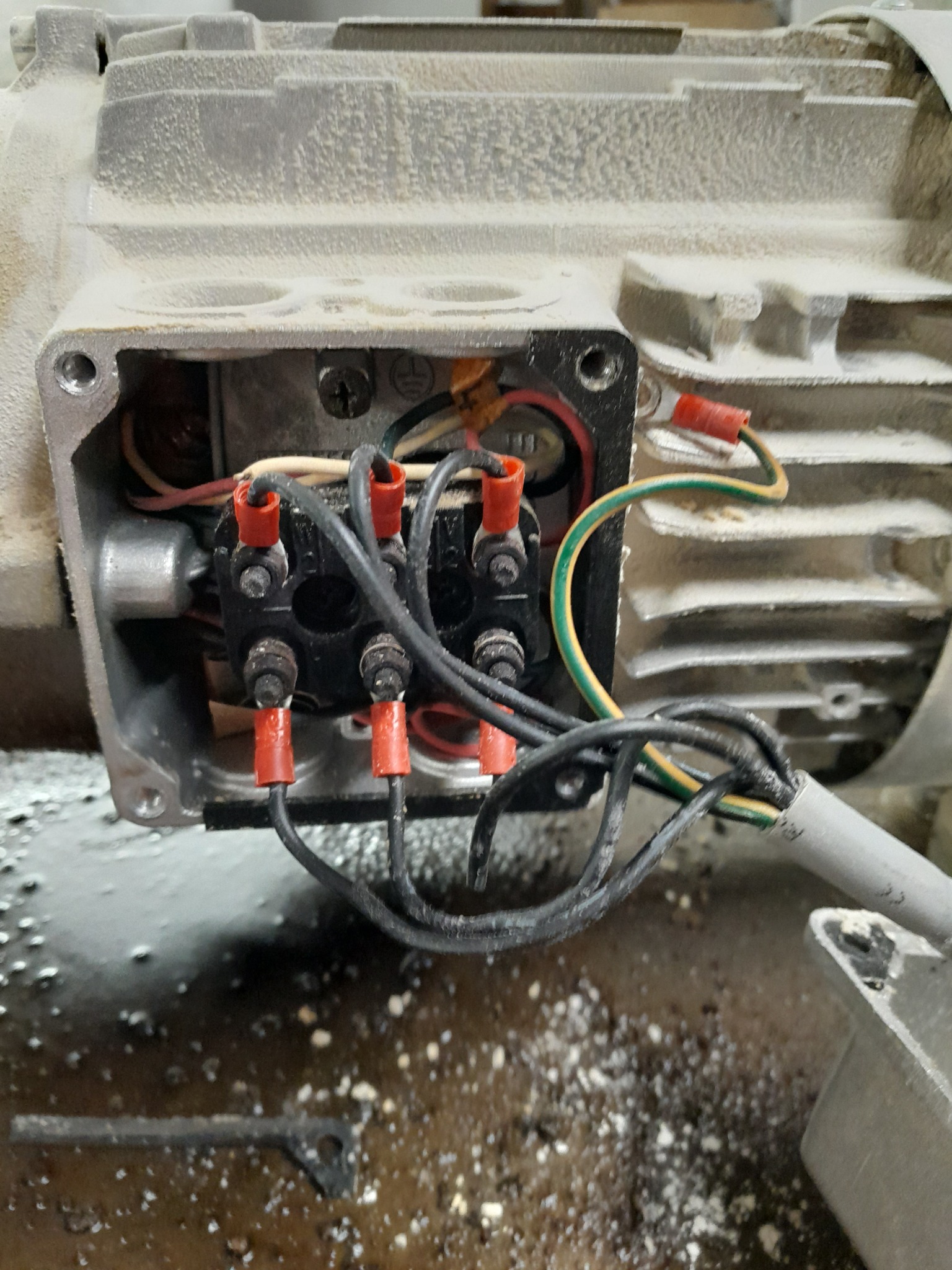 Otwarta puszka przyłączeniowa silnika elektrycznego z widocznymi przewodami zakończonymi czerwonymi konektorami oczkowymi, podłączenie do listwy zaciskowej z czarnymi śrubami, zakurzona obudowa...