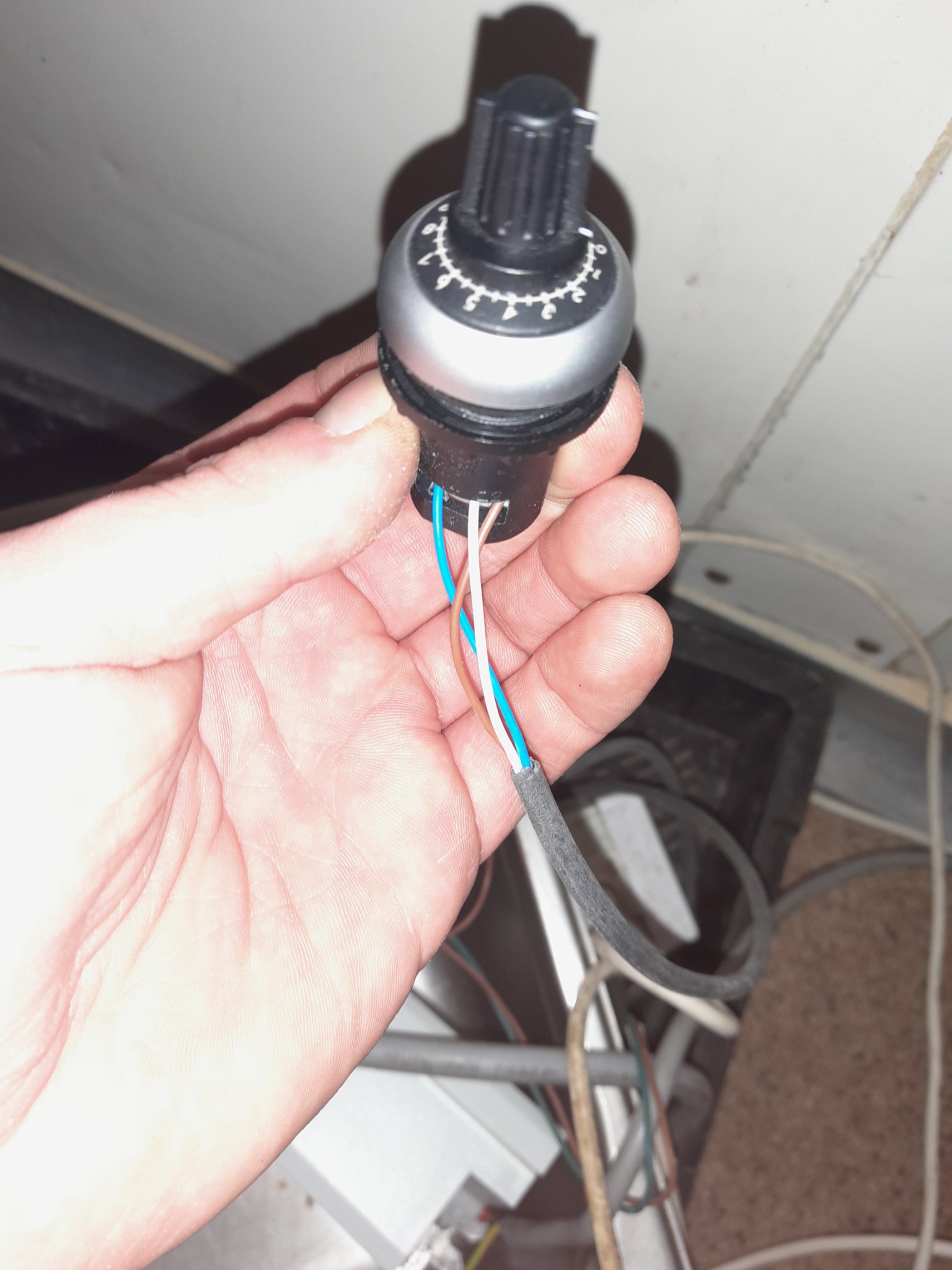Dłoń trzyma pokrętło regulatora z podłączonymi przewodami (niebieski, biały, brązowy) na tle instalacji elektrycznej.
