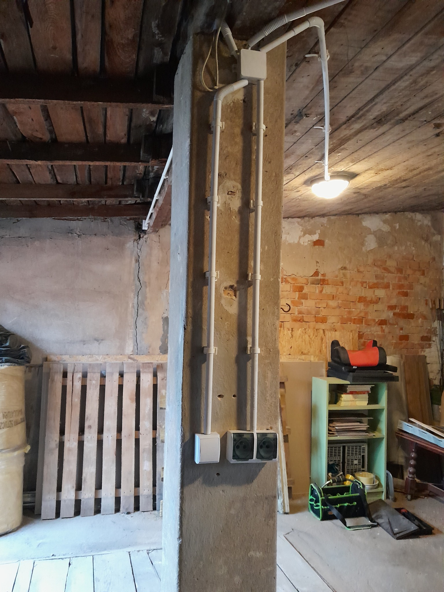 Betonowy słup z zamontowanymi gniazdkami elektrycznymi i białymi rurami instalacyjnymi w surowym wnętrzu z drewnianym stropem i ceglaną ścianą w tle. Widoczna lampa sufitowa i narzędzia.