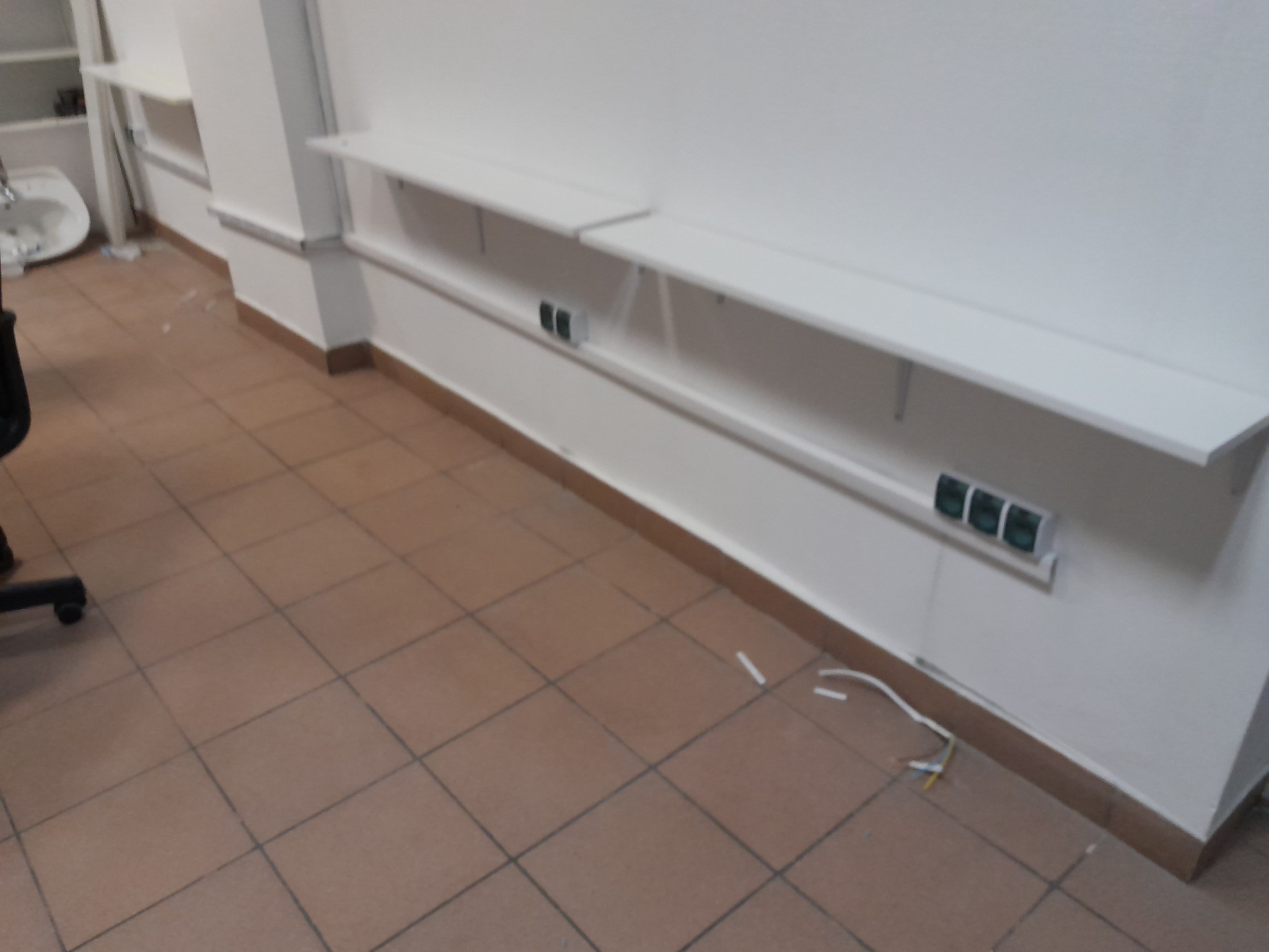 Białe półki zamontowane na ścianie z widocznymi gniazdkami elektrycznymi, kable zwisają na brązowej podłodze z płytek.