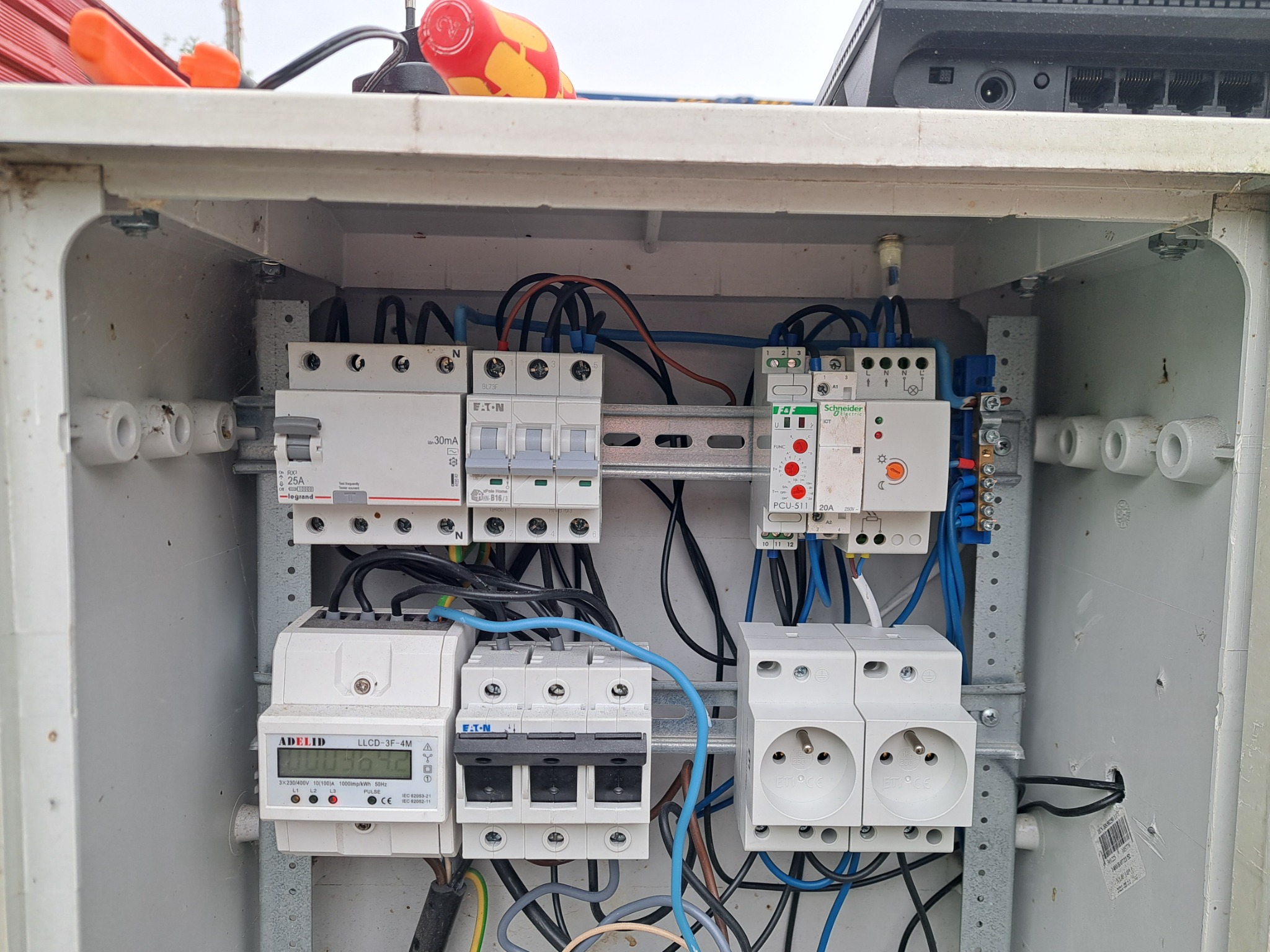Wnętrze skrzynki elektrycznej z zamontowanymi bezpiecznikami EATON, licznikiem ADELID, gniazdkami ETI i modułem Schneider Electric, widoczne przewody w kolorach niebieskim, czarnym i brązowym.