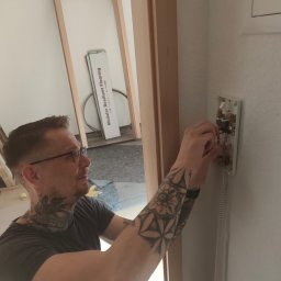 Goldfinger - Elektryk z tatuażami naprawia domofon na białej ścianie. Widoczne przewody i elementy elektroniki. W tle materiały budowlane.