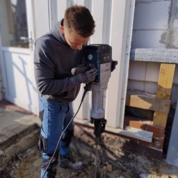 Goldfinger - Pracownik w szarej bluzie obsługuje młot udarowy Bosch GSI 7 VC, przygotowując wykop pod instalację przy białej ścianie budynku z pustaków.